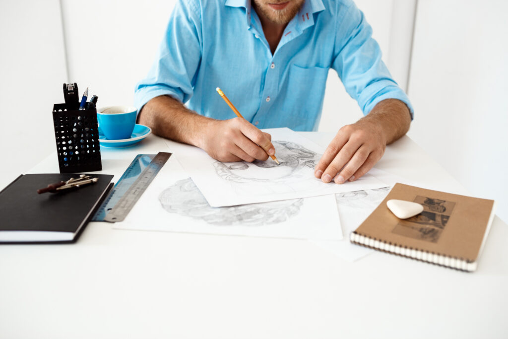 Hands of young businessman sitting at table with pencil drawing portrait. White modern office interior background.